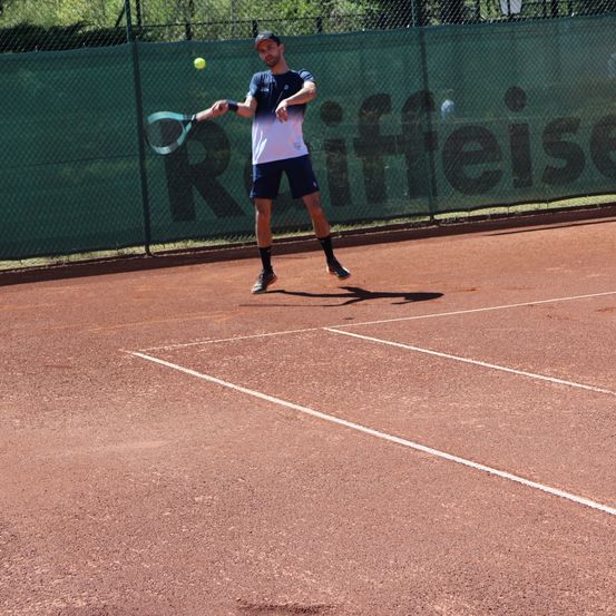 Ein Mann spielt Tennis auf einem Platz. Er schwingt seinen Schläger, um einen Ball in der Luft zu treffen. Der Platz ist mit weißen Linien markiert.