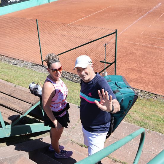 Ein Mann und eine Frau gehen die Stufen hinauf, der Mann winkt und sie befinden sich in der Nähe eines Tennisplatzes.