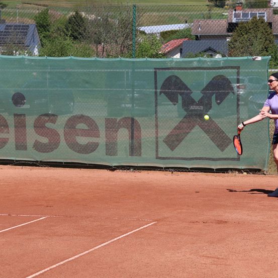 Eine Frau spielt Tennis auf einem Sandplatz. Hinter ihr zeigt ein grünes Netz das Wort 'eisen' und ein Logo. Im Hintergrund befinden sich Häuser mit Solarpanels.