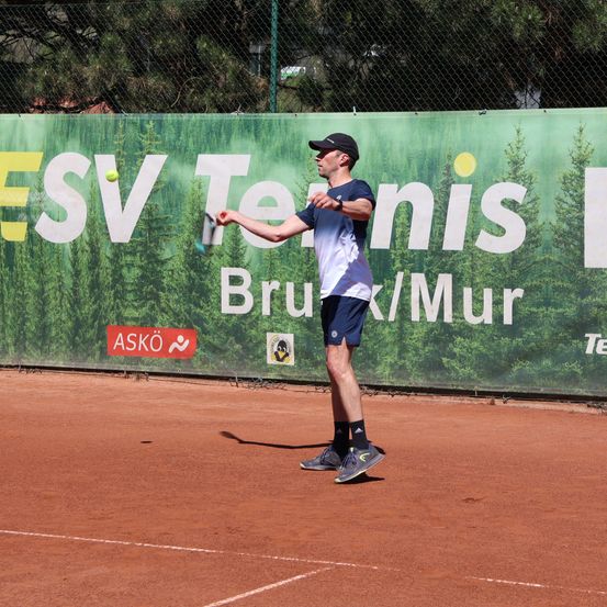 Ein Mann schwingt einen Tennisschläger auf einem Tennisplatz, mit einer grünen Wand im Hintergrund, auf der in großen weißen Buchstaben ESV Tennis steht.