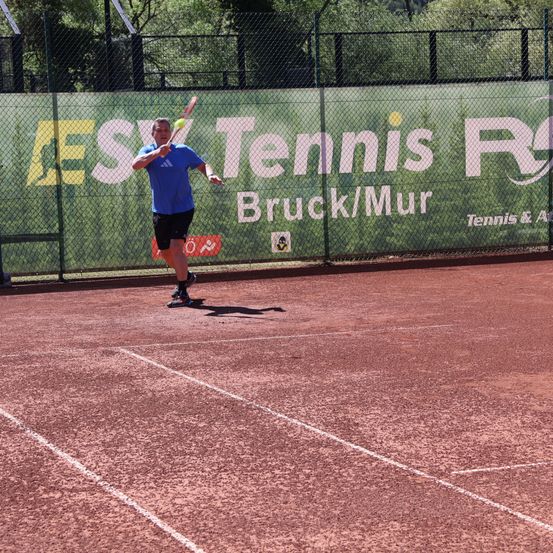 Ein Mann in einem blauen Shirt und schwarzen Shorts spielt Tennis auf einem Platz mit einem grünen Zaun. Der Zaun hat ein Banner, das für ES Tennis Bruck/Mur wirbt.