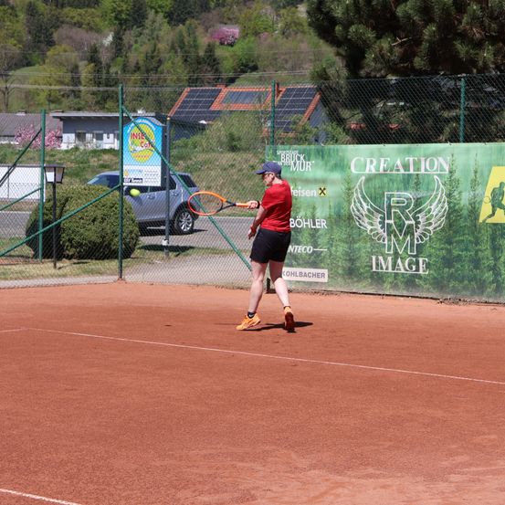Ein Mann in einem roten Shirt und schwarzen Shorts spielt Tennis auf einem Sandplatz und trifft einen gelben Ball mit einem Tennisschläger.