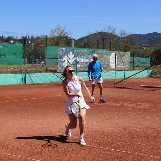 Zwei Personen spielen Tennis auf einem Freiluftplatz, eine Frau in einem weißen Kleid und ein Mann in Blau. Das Spielfeld ist von Zäunen umgeben, im Hintergrund befindet sich ein grüner Banner.