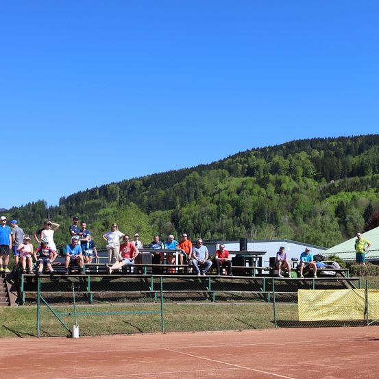 Eine Gruppe von Menschen sitzt auf Tribünen eines Tennisplatzes mit einem grünen Zaun und einem klaren blauen Himmel. Sie tragen Mützen und Turnschuhe. Eine Person in einem gelben Shirt steht auf der rechten Seite. Der Platz hat eine weiße Linie. In der Ferne gibt es einen Berg mit Bäumen.