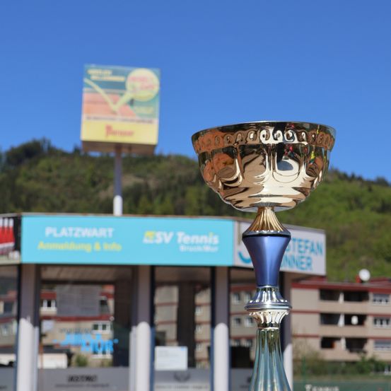 Ein goldener Pokal steht vor einem blauen Schild, wahrscheinlich bei einem Tennis-Event. Dahinter befindet sich ein Gebäude mit einem roten Dach und Bäumen. Der Himmel ist blau.