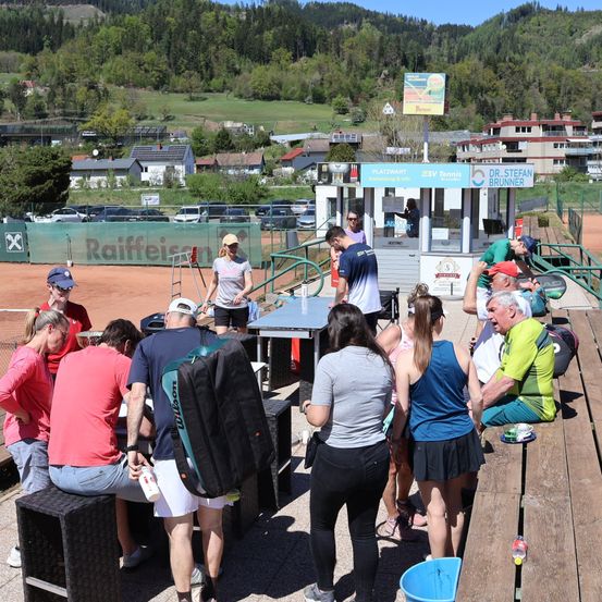 Eine Gruppe von Tennisspielern und Zuschauern befindet sich auf einem Tennisplatz, einige sitzen auf Bänken und andere stehen. Eine Person lehnt sich an eine Metallbarriere.