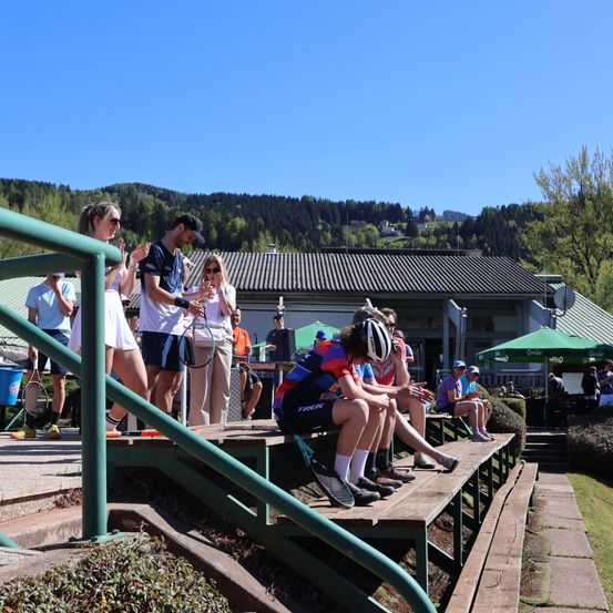 Eine Gruppe von Menschen sitzt auf Tribünen, einige halten Tennisschläger in der Hand, unter einem klaren blauen Himmel, mit Bergen und Grün im Hintergrund.