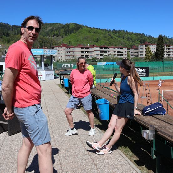 Eine Gruppe von Menschen steht neben einem Tennisplatz. Ein erwachsener Mann hält ein Mikrofon, während eine erwachsene Frau auf einer Bank steht und einen Tennisschläger in der Hand hält.