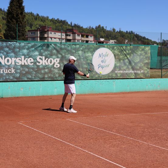 Ein Mann in einem blauen Shirt spielt Tennis auf einem Sandplatz. Der Platz hat grüne Wände und ein großes Banner.