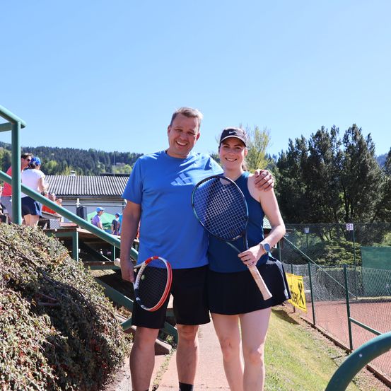 Ein Mann und eine Frau stehen auf einem Tennisplatz, lächeln und posieren für ein Foto. Der Mann trägt ein blaues T-Shirt, während die Frau eine Kappe und ein blaues ärmelloses Top trägt. Sie halten Tennisschläger und stehen vor einer Kulisse aus Bäumen und einem Zaun.