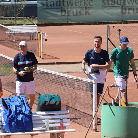 Drei Männer befinden sich auf einem Tennisplatz. Einer hält einen Tennisschläger und einen Ball. Ein anderer hält einen Hut. Der dritte hat eine Wasserflasche. Sporttaschen und Ausrüstung sind in der Nähe. Eine Bank ist im Hintergrund.