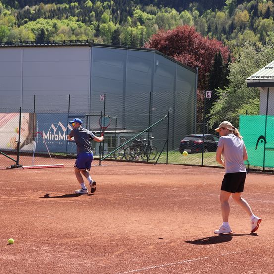 Zwei Personen spielen Tennis auf einem Außenplatz. Einer ist dabei, einen Ball in der Luft zu schlagen. In der Nähe befindet sich ein Gebäude mit einem Schild und geparkten Autos.