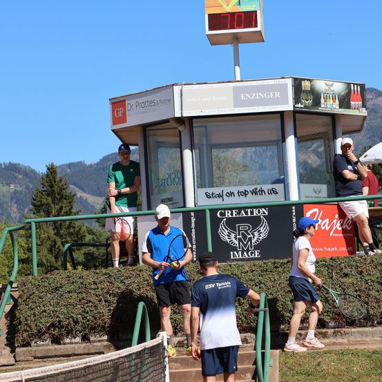 Eine Gruppe von Tennisspielern versammelt sich auf einem Tennisplatz. Die Temperatur beträgt 70 Grad. Ein Spieler mit einer blauen Kappe hält einen Tennisschläger.