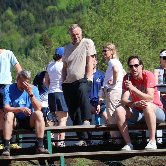 Eine Gruppe von Menschen sitzt draußen auf einer Bank. Ein Mann mit Sonnenbrille hält ein Mikrofon. Ein anderer Mann steht mit den Händen in den Hüften. Die Leute halten Tennisschläger.