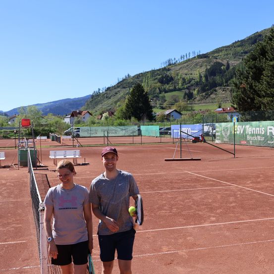 Zwei Personen stehen auf einem Tennisplatz mit einem Berg im Hintergrund. Beide halten Tennisschläger. Der Platz hat grüne Banner mit Text.