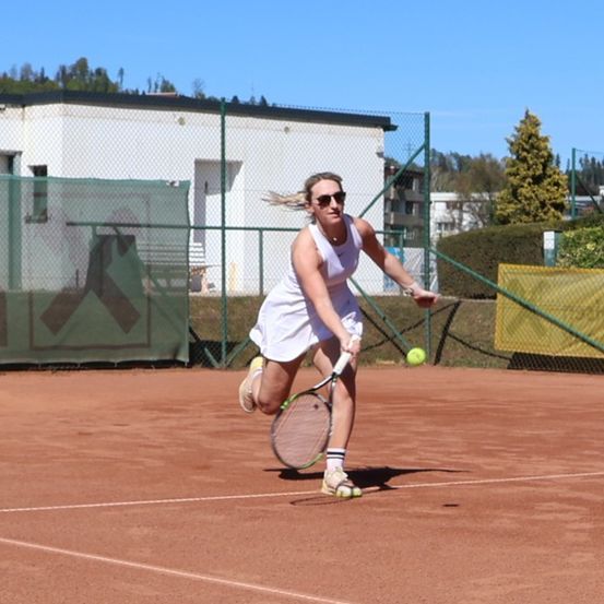 Eine Frau in einem weißen Kleid und Sonnenbrille spielt Tennis auf einem Sandplatz. Sie ist bereit, einen gelben Ball mit ihrem Schläger zu schlagen.