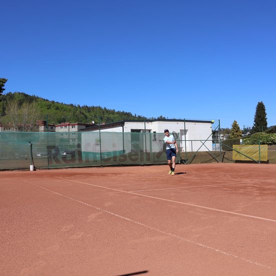 Ein Mann in blauen Shorts und einem Hut läuft auf einem roten Tennisplatz. Im Hintergrund befindet sich ein Zaun mit einem Banner, das Rainier besagt.