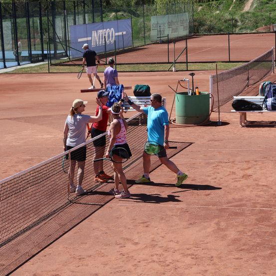 Eine Gruppe von Tennisspielern schüttelt sich über das Netz die Hände, während andere zuschauen und einer zur Seite geht. Der Platz ist von einem Zaun umgeben und hat einen großen grünen Wasserbehälter.