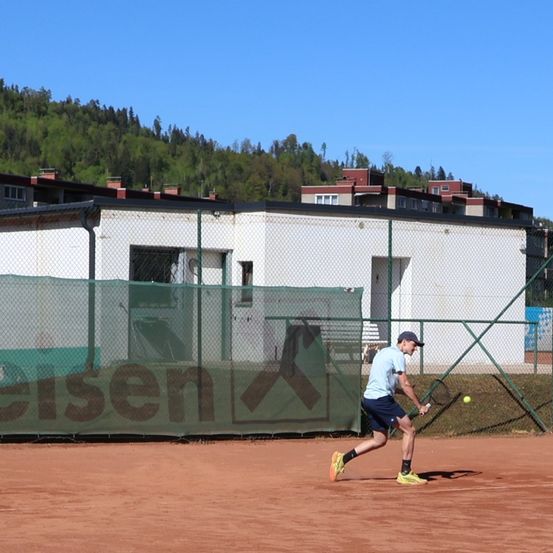 Ein Mann schwingt einen Tennisschläger auf einem Platz, im Hintergrund steht ein Gebäude. Das Gebäude hat einen Zaun mit einem Schild für cisen.