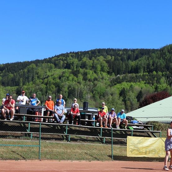 Zuschauer sitzen auf Tribünen unter blauem Himmel und beobachten ein Tennisspiel. Ein grüner Zaun und ein gelbes Banner sind im Vordergrund.