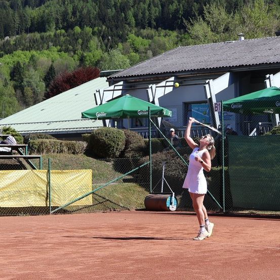 Eine Tennisspielerin in weißem Kleid und Schuhen, die einen Schläger hält, schlägt einen Ball auf einem Tennisplatz. Hinter ihr befindet sich ein Gebäude mit grünen Sonnenschirmen.