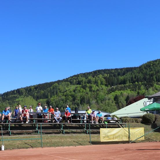 Eine Gruppe von Menschen sitzt auf der Bank eines Tennisplatzes. Sie scheinen ein Spiel zu beobachten. Der Platz ist von einem Zaun umgeben.