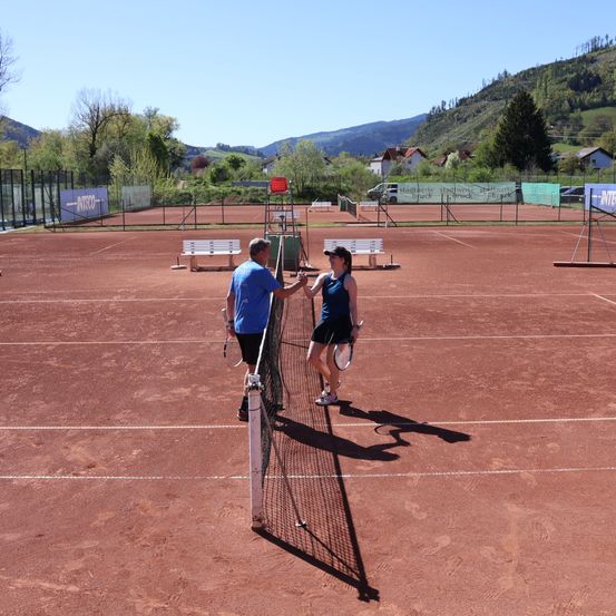 Ein Mann und eine Frau schütteln sich die Hände auf einem Tennisplatz. Sie halten Tennisschläger in den Händen. Der Platz ist von einem Zaun umgeben, und es gibt Bänke und einen Berg im Hintergrund.
