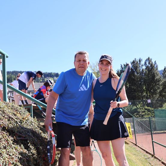 Ein älterer Mann und eine junge Frau halten Tennisschläger in der Hand und lächeln für ein Foto auf einem Tennisplatz. Dahinter befinden sich Personen auf Treppen und in der Nähe von Bäumen.