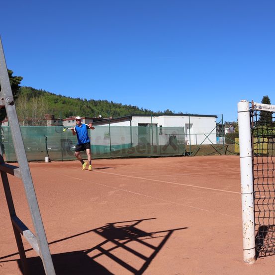 Ein Tennisspieler in einem blauen Shirt und schwarzen Shorts schlägt einen Tennisball auf einem Freiluftplatz.