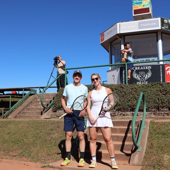 Zwei Tennisspieler posieren für ein Foto auf einem Tennisplatz mit einem Fotografen und einem Mann in einer Kabine im Hintergrund.