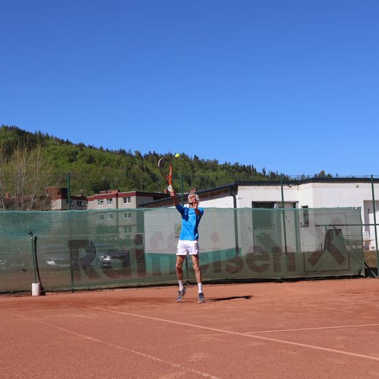 Ein Mann in einem blauen Shirt und weißen Shorts spielt Tennis auf einem Platz. Er hält einen Tennisschläger und springt, um den Ball zu schlagen. Dahinter befindet sich ein Zaun mit einer großen Werbung für Raiffeisen.