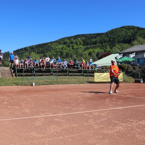 Ein Tennisspieler übt auf einem Sandplatz. Zuschauer sitzen auf den Tribünen. Im Hintergrund befinden sich Berge und Grün.