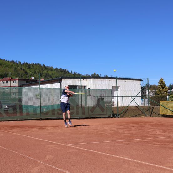 Ein Tennisspieler springt auf einem Tennisplatz, um einen Ball zu schlagen. Hinter ihm steht ein weißes Gebäude mit Zaun.