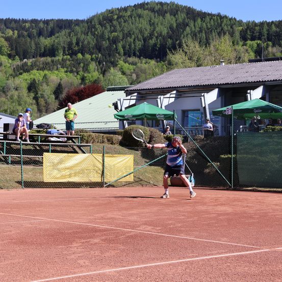 Ein Tennisspieler schlägt einen Ball auf einem Sandplatz, während Zuschauer von den Tribünen zuschauen und grüne Sonnenschirme in der Nähe stehen.