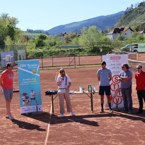 Fünf Personen stehen auf einem Tennisplatz. Eine Frau spricht in ein Mikrofon auf einem Tisch. Plakate sind hinter ihr und neben ihr. Ein Mann steht neben dem Plakat, und eine Bergkette ist im Hintergrund.