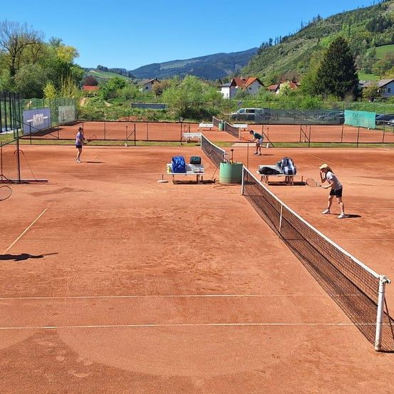 Ein Tennisplatz mit zwei Spielern im Hintergrund, umgeben von einem Zaun und einer Berglandschaft.