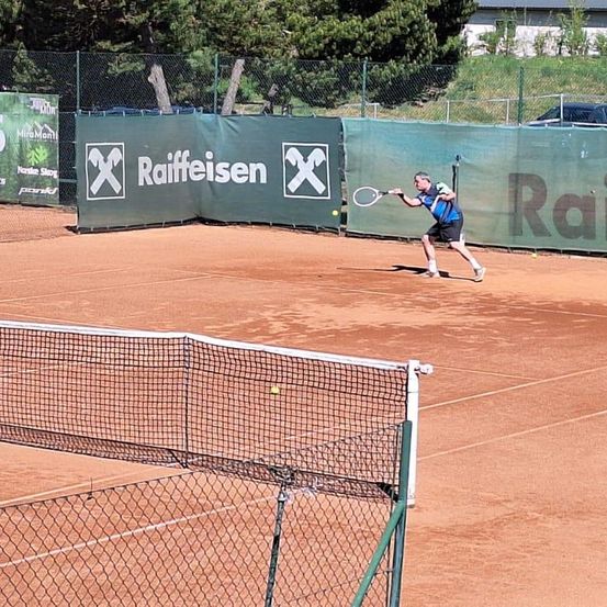 Ein Mann in einem blauen Shirt und schwarzen Shorts spielt Tennis auf einem Sandplatz. Der Platz hat grüne Banner mit Logos und dem Wort Raiffeisen. Bäume und ein Zaun sind im Hintergrund.
