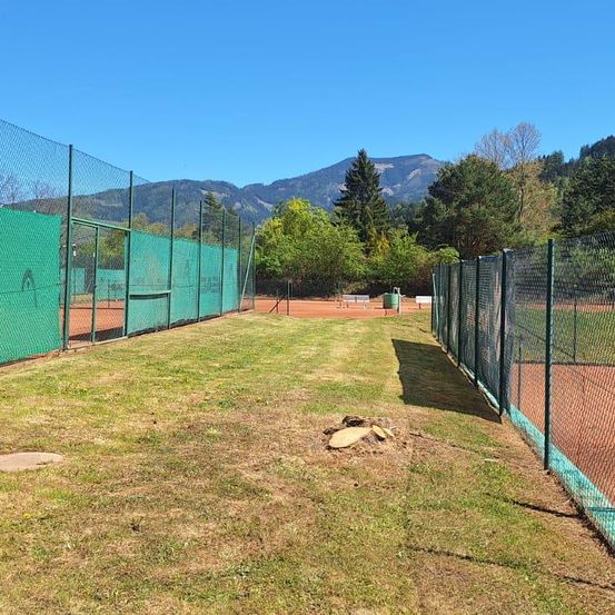 Ein Tennisplatz umgeben von grünen Netzen und einem Rasenfeld. Im Hintergrund befinden sich Berge und Bäume.
