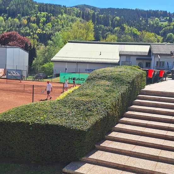 Ein Tennisplatz mit Heckenbarriere, zwei Spielern und einem Gebäude mit grünem Banner. Ein Berg mit Bäumen im Hintergrund.