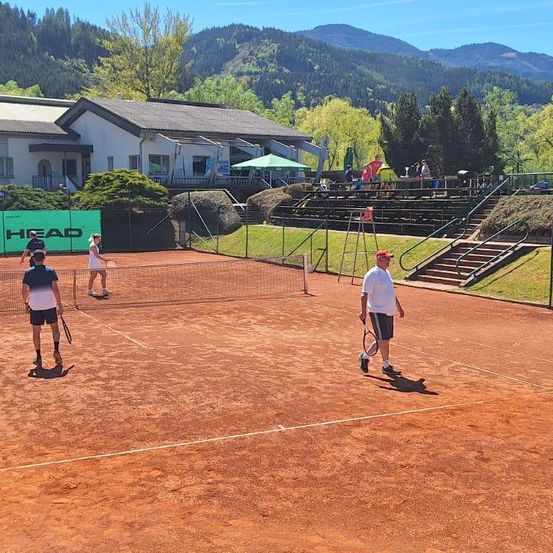 Ein Tennisplatz mit Spielern und einem Zuschauer, mit einem Gebäude und Bergen im Hintergrund.