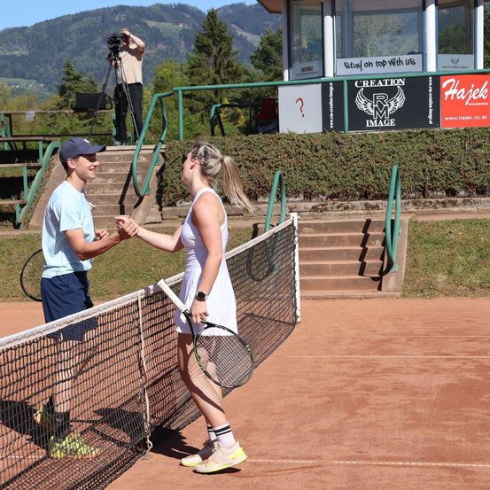Auf einem Tennisplatz schütteln sich zwei Spieler, ein Mann und eine Frau, die Hände. Dahinter hält ein Kameramann den Moment fest. In der Nähe führen Stufen zu einer Struktur mit einem Schild, auf dem 'CREATION IMAGE' steht. Bäume und Berge sind im Hintergrund.