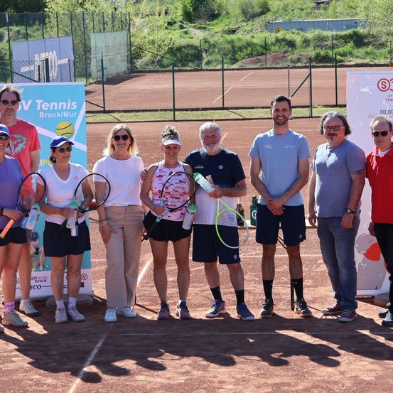 Gruppe von Menschen auf einem Tennisplatz, die Tennisschläger halten. Zwei Banner sind im Hintergrund.