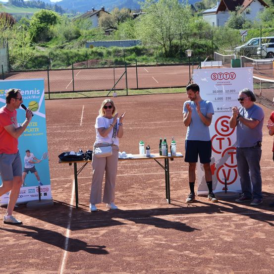 Ein Tennisevent mit vier Personen auf einem Platz, einer mit Mikrofon, Flaschen auf einem Tisch und Bannern an den Seiten.