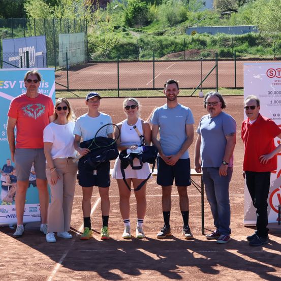 Eine Gruppe von Menschen, wahrscheinlich ein Tennisteam, posiert für ein Foto auf einem Tennisplatz. Sie tragen alle Turnschuhe und einige halten Tennisschläger. Es gibt zwei Banner an der Seite des Platzes.