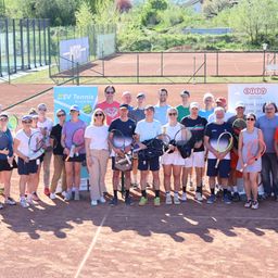 Eine Gruppe von Tennisspielern auf einem Tennisplatz, mit Schlägern, in Reihen stehend, mit einem Banner und Zaun im Hintergrund.