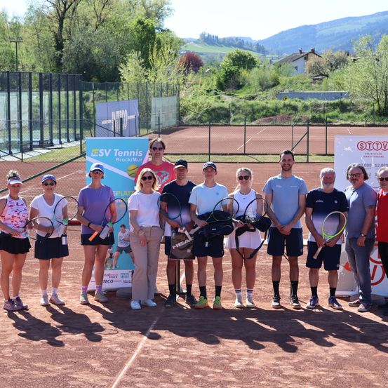 Eine Gruppe von Tennisspielern und Trainern posiert auf einem Tennisplatz mit einem Hintergrund eines Tennisclub-Banners. Bäume und Berge sind im Hintergrund.