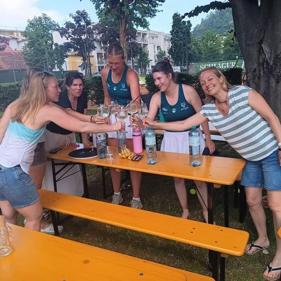 Eine Gruppe von Frauen in Sportkleidung stoßen mit Wassergläsern an einem Picknicktisch im Freien an, mit Bäumen und Gebäuden im Hintergrund.