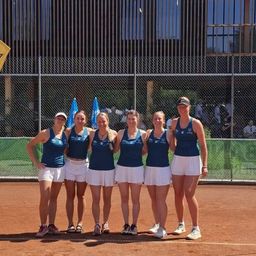 Eine Gruppe von Tennisspielerinnen posiert auf einem Tennisplatz für ein Foto. Sie tragen passende Uniformen. Hinter ihnen steht ein Zaun mit Flaggen.