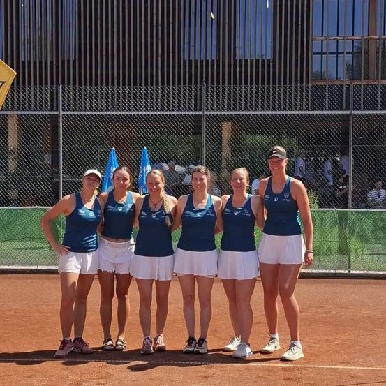 Eine Gruppe von Tennisspielerinnen posiert auf einem Tennisplatz für ein Foto. Sie tragen passende Uniformen. Hinter ihnen steht ein Zaun mit Flaggen.