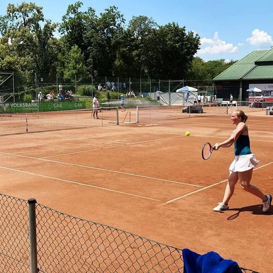 Eine Frau schwingt einen Tennisschläger auf einem Sandplatz, eine gelbe Kugel ist in der Luft. Auf der gegenüberliegenden Seite des Platzes spielen Leute Tennis.